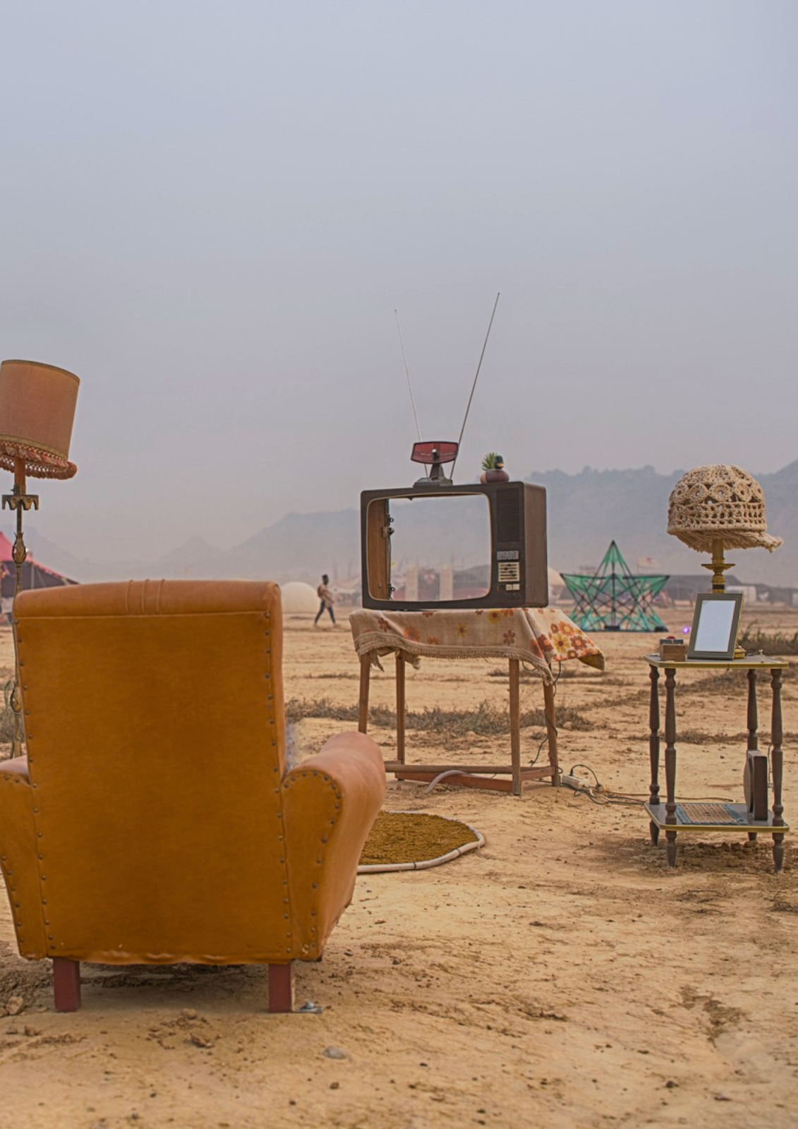 A surreal desert living room — a vintage armchair, an old TV, a lamp, and scattered objects creating an unexpected domestic scene on the playa
