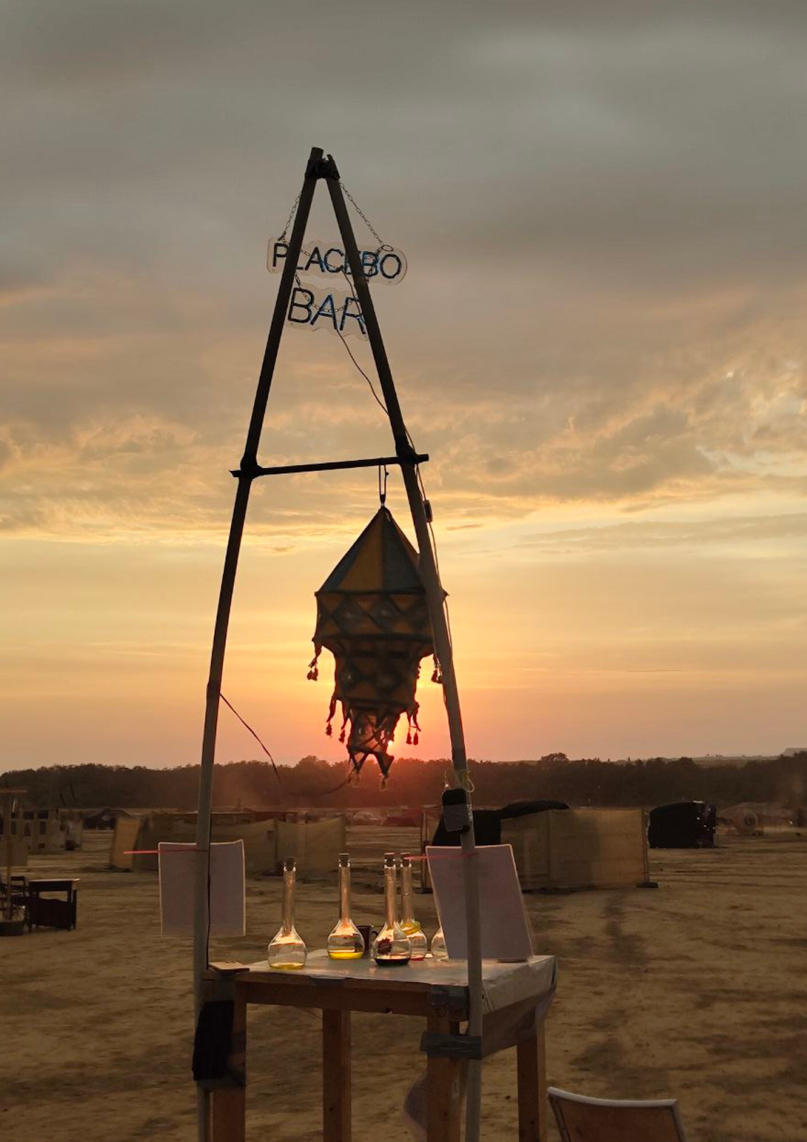 The Placebo Bar — a handcrafted desert installation with a hanging lantern, glass bottles, and a sunset glow over the playa